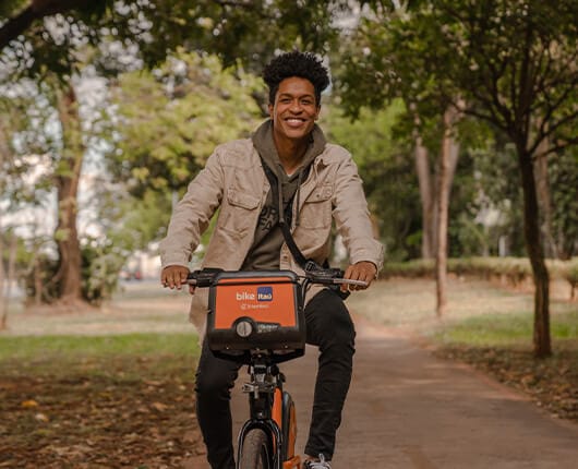 Homem de casaco bege pedalando a bike Itaú no parque.